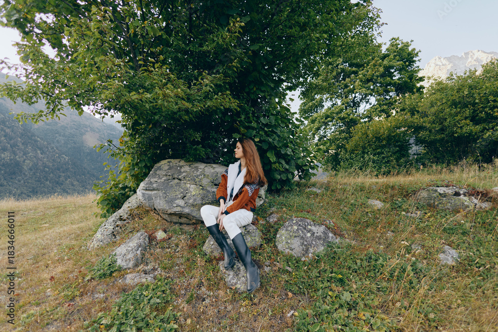Obraz premium Woman sitting on rock under tree in mountain meadow outdoors, portrait of a traveler in boots and warm coat enjoying the landscape and peaceful autumn nature with distant peaks.