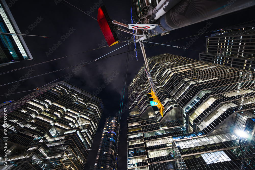 Fototapeta premium Night reflections rise into a maze of glass and steel as a glowing traffic light hangs beneath towering skyscrapers. The scene feels like a futuristic canyon where geometry, light