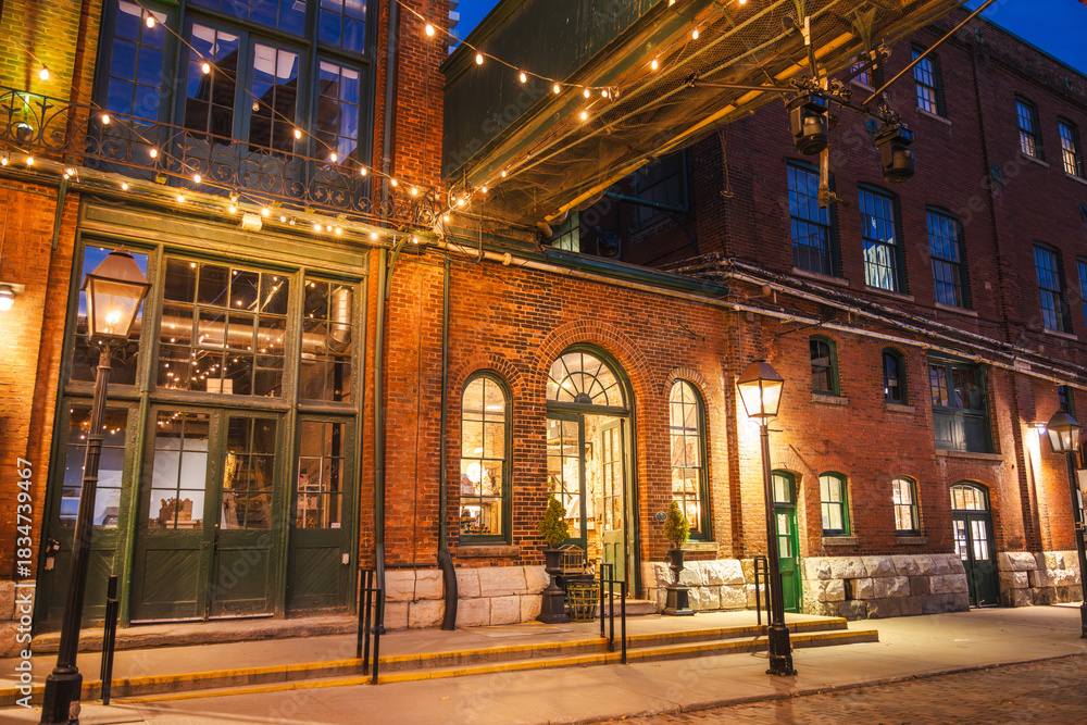 Obraz premium Historic brick architecture illuminated by warm streetlights and string lights in the Distillery District of Toronto at night.