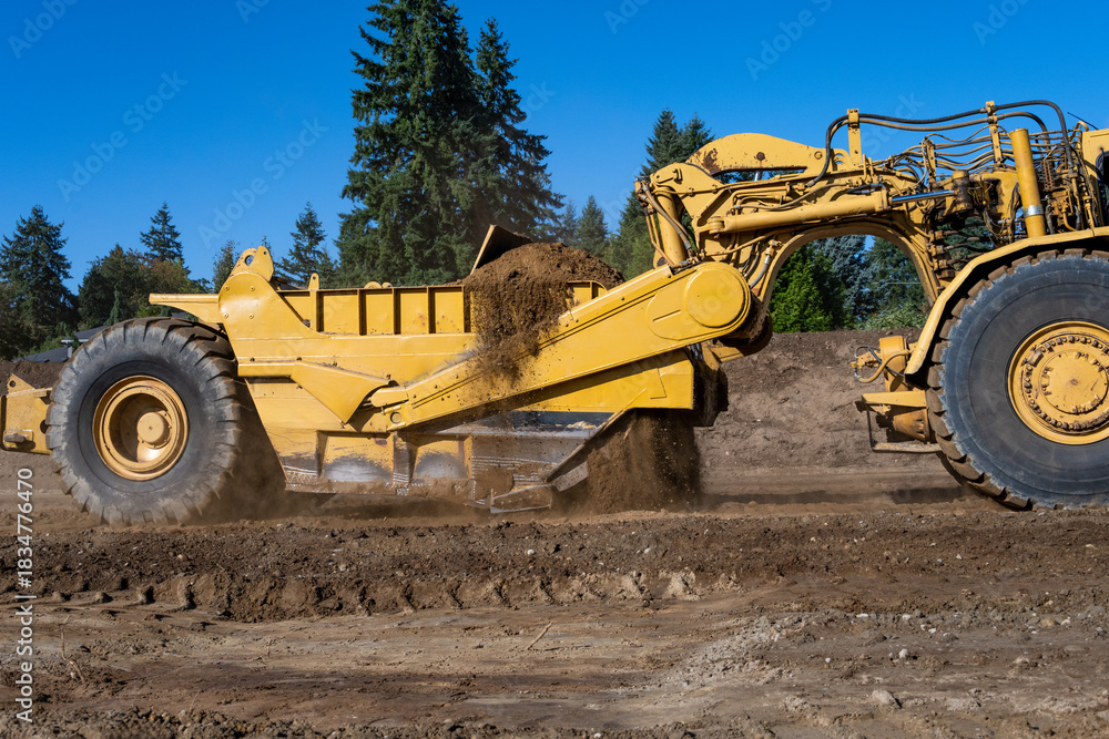 Fototapeta premium Wheel Tractor Scraper moving mineral dirt and working on groundwork for new housing development project, leveling ground for road works, as a construction background 