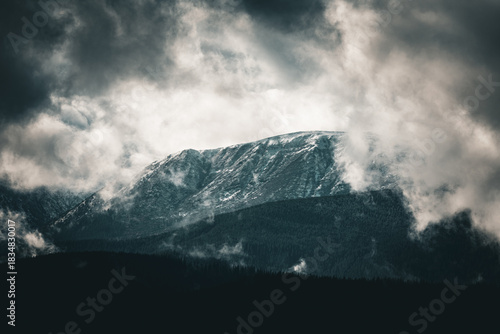 Moody, cool-toned clouds encircle a snowcapped mountain ridge above dark evergreen layers. Mist hangs over the forested slopes, creating a cinematic, alpine panorama with bold contrast and depth.
