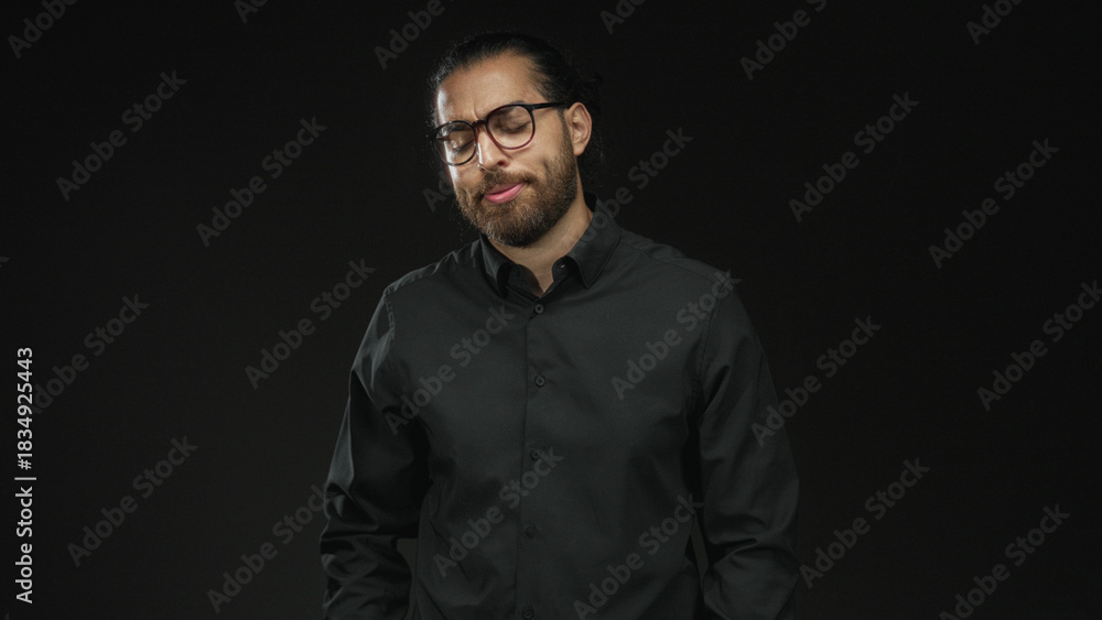 Obraz premium Man with glasses and beard, eyes closed and head tilted, hands in pockets wearing a dark shirt in a black studio; contemplative.