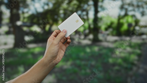 Man holding white credit card in outdoor park setting with lush green background, showcasing finance and technology concept in a natural environment.