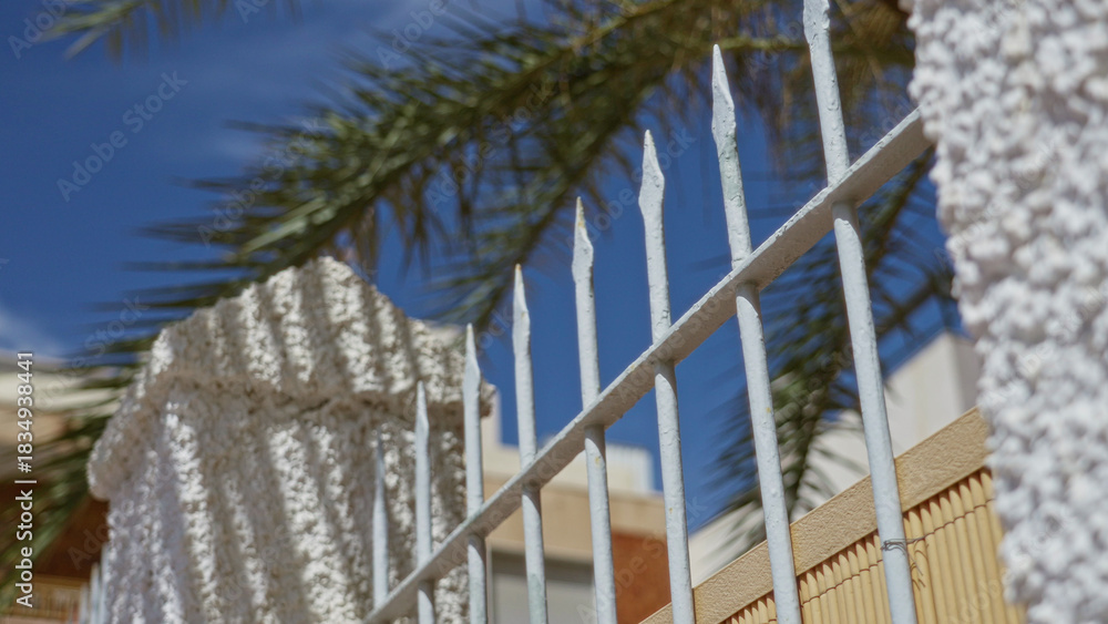 Naklejka premium Sunny outdoor scene of a metal gate and textured house wall framed by palm leaves under a clear blue sky.