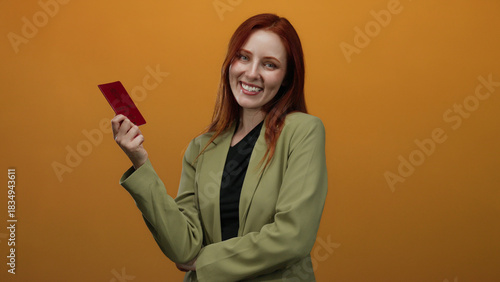Schilderij op canvas Woman with red hair joyfully holds a norwegian passport against an isolated orange background, highlighting personal identity and global travel