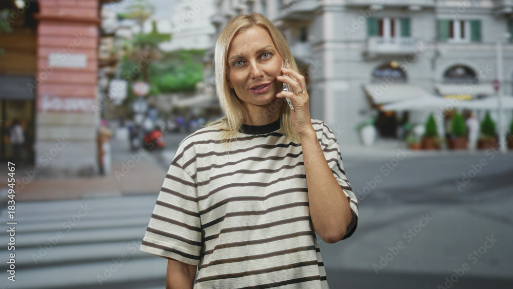 Fototapeta premium Woman holds phone to ear at street crosswalk near cafe awnings and city buildings, striped shirt visible; calm conversation.