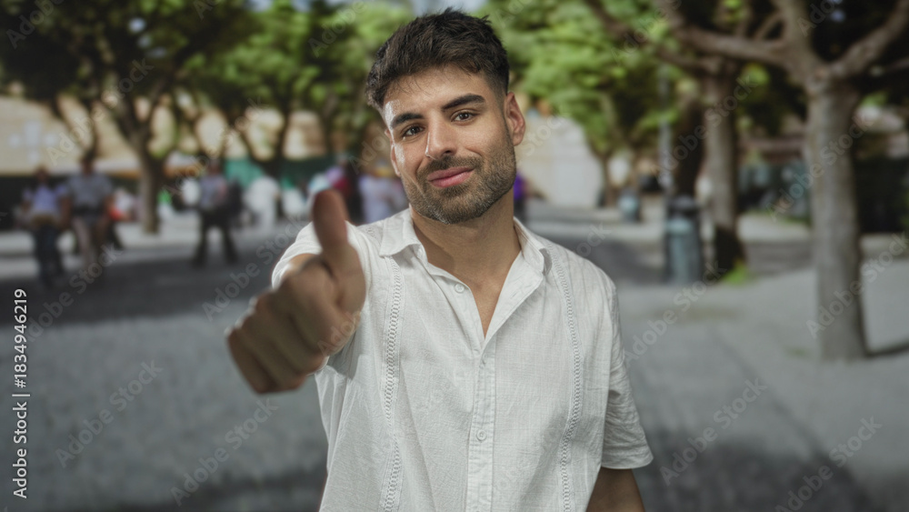 Naklejka premium Confident man makes vigorous thumbs up gesture with bare thumb on street amid blurred pedestrians; approval.
