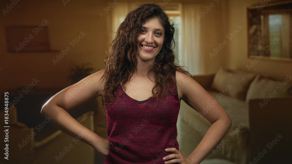Fototapeta premium Woman with hands on hips and bare arms visible in a living room inside a building, smiling at camera; confidence comfort.