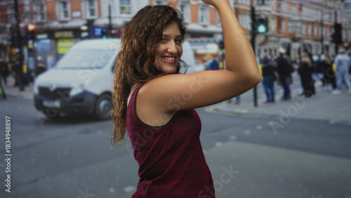 Schilderij op canvas Young woman flexes bare upper arm and smiles while posing amid a busy city street with pedestrians, cars and shopfronts visible; confidence