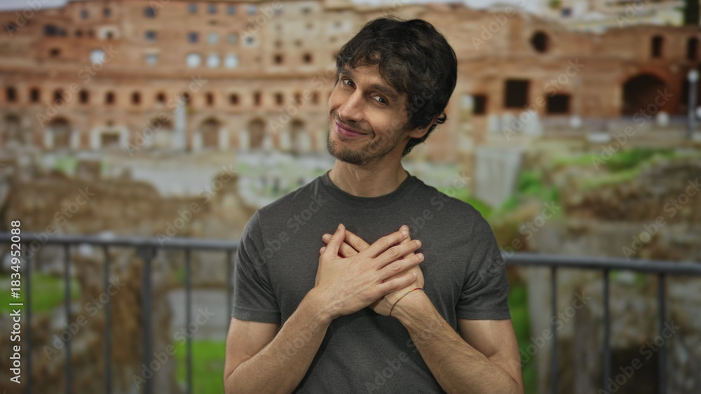 Fototapeta premium Man in casual grey shirt touches chest and looks at camera amid ancient roman ruins at building; sincerity.
