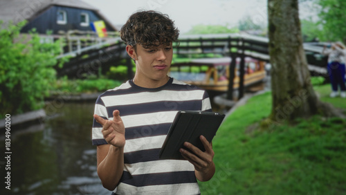 Teen man holds tablet with one hand and presses palm to chest while reading a startling notification on a canal street with wooden bridge nearby; surprise reflection.