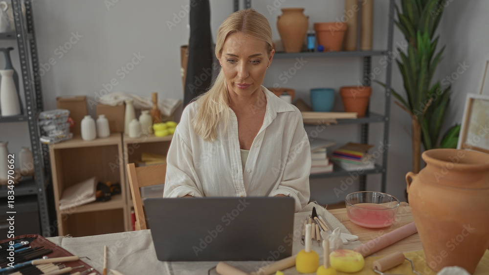 Fototapeta premium Woman types on laptop surrounded by pottery tools and clay jars under natural light in studio; creative concentration.