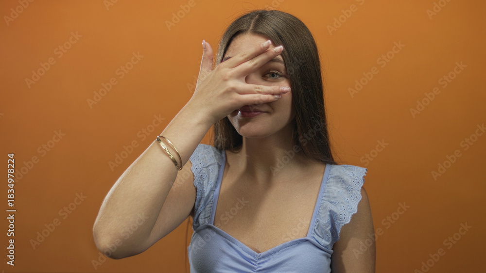 Fototapeta premium Teenage girl covers eyes with hand in studio against orange wall; vulnerability introspection shyness.
