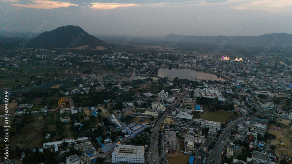 Fototapeta premium Aerial view of Pushkar Lake during the fair, capturing cloud reflections on the water, glowing roller-coaster lights, lakeside houses and ghats, with a warm sunset over the Aravali hills in the back
