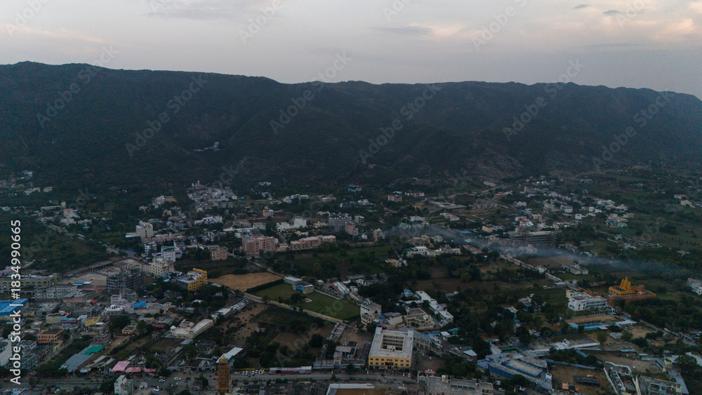 Obraz premium Aerial pan shot of Pushkar town at sunset, showing houses, Pushkar Lake, glowing evening light, cloudy sky, and the Aravali hills creating a scenic backdrop across the serene landscape from above.