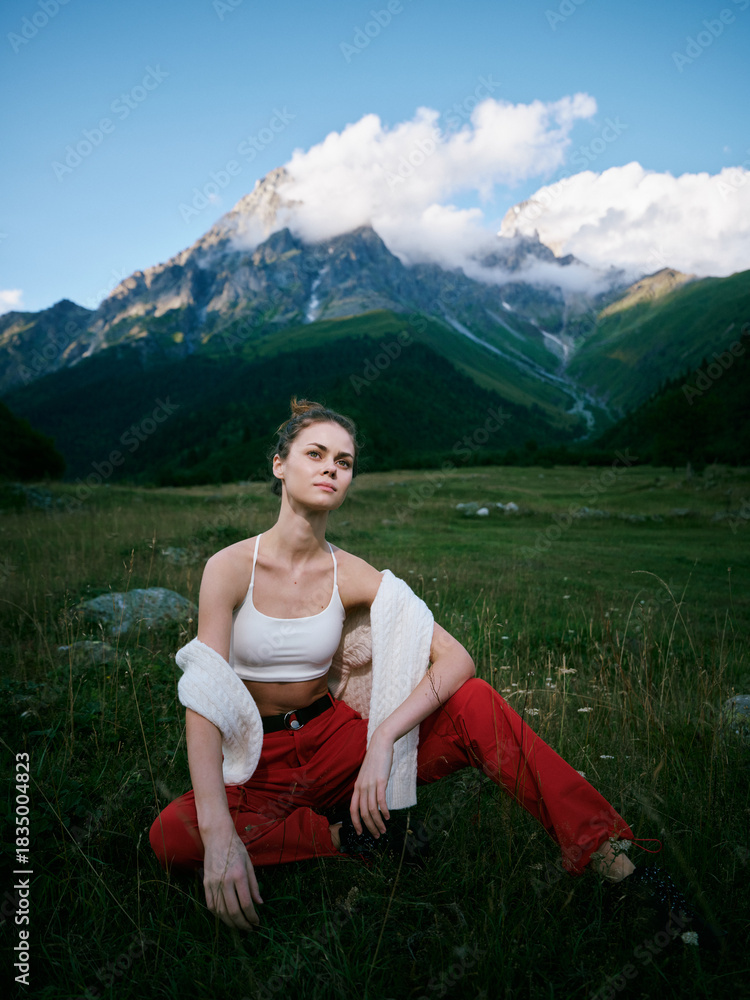 Fototapeta premium Woman sits in a grassy field with a mountain backdrop, wearing a white crop top and red pants, relaxed pose in natural light, outdoor scenery and serene alpine landscape