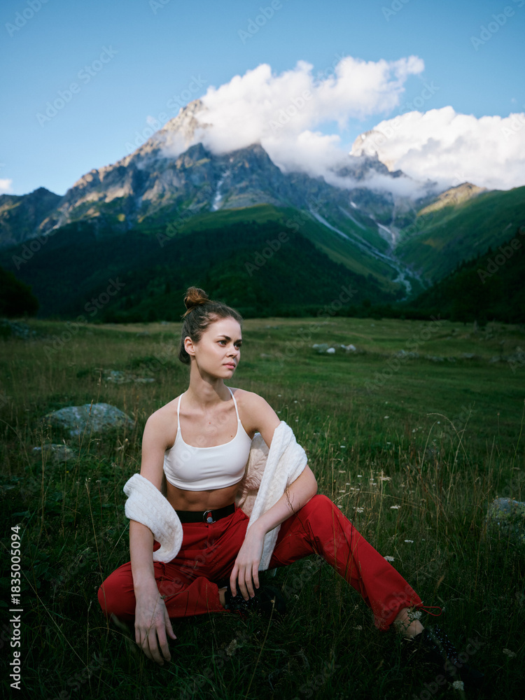 Obraz premium Mountain meadow scene with a stylish woman sitting in green grass, white crop top, red pants, relaxed pose, dramatic clouds over distant peaks, wildflowers around, tranquil alpine atmosphere and