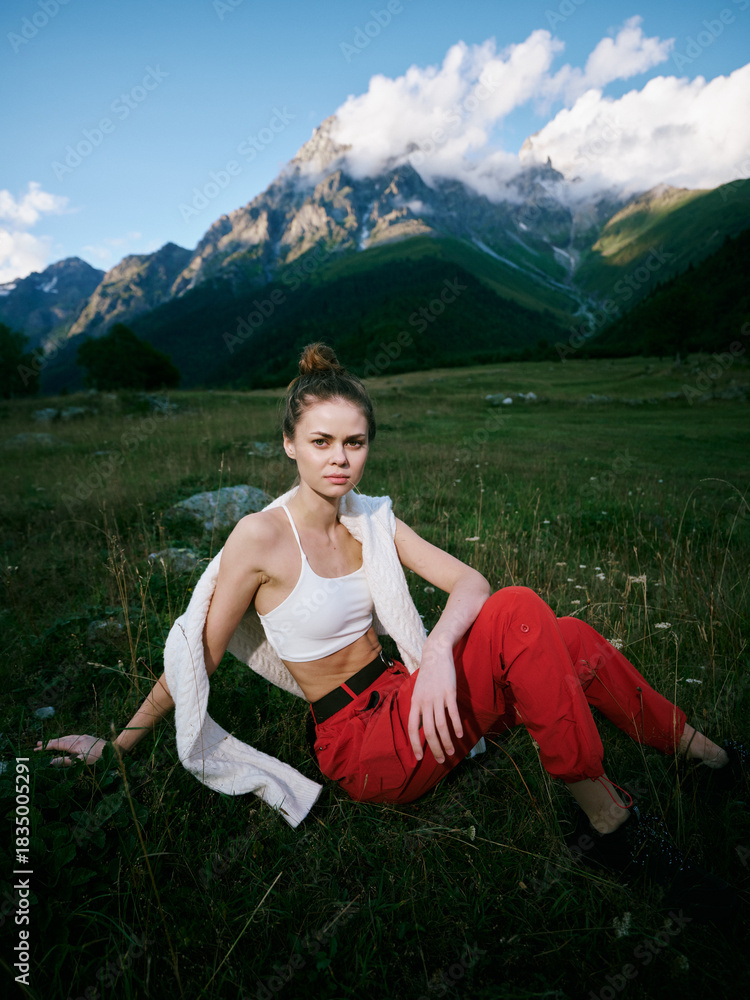 Fototapeta premium Woman in white top and red pants sits in a meadow with towering mountains and clouds, outdoor fashion and nature inspiration for travel, leisure, and adventure photography