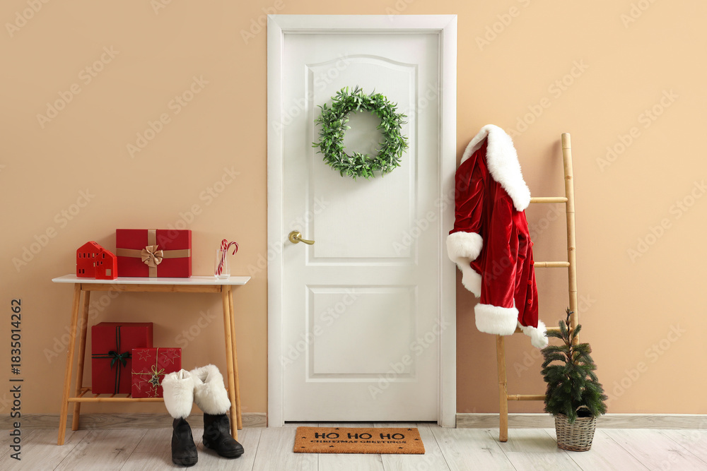 Fototapeta premium Christmas home entrance with white door, green wreath, Santa costume on ladder, gift boxes, boots and festive decor in cozy minimalist interior