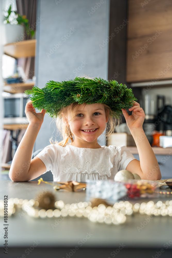 Naklejka premium Little girl wearing a Christmas wreath on her head and laughing during holiday decorating. Festive joy, family traditions, and playful moments during Christmas preparation.
