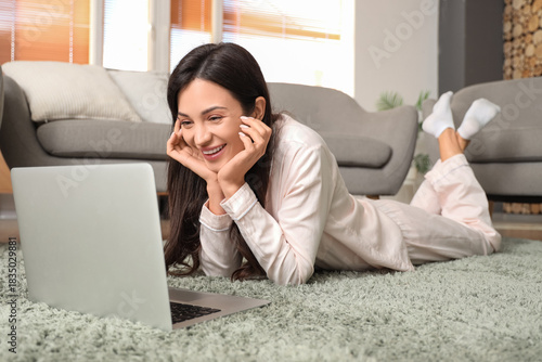 Smiling young woman in pajamas lying on carpet and using laptop at home, rela...