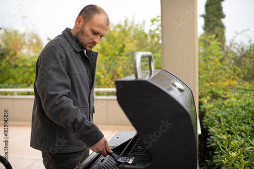 Man inspecting a barbecue grill on a sunny terrace, preparing food for outdoor cooking, home leisure, summer lifestyle.