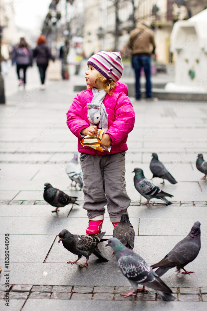 Obraz premium Little girl feeding pigeons on city street in winter clothes
