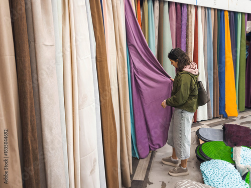 Woman choosing curtains in textile store, comparing colors and textures while holding purple drapery fabric. Bright rows of curtain materials create vivid shopping atmosphere and home decor context.