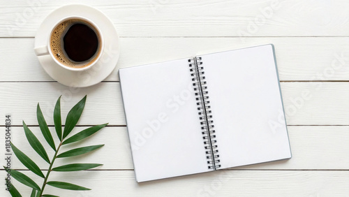 Coffee cup and blank notebook placed on a wood table with a green leaf on the side