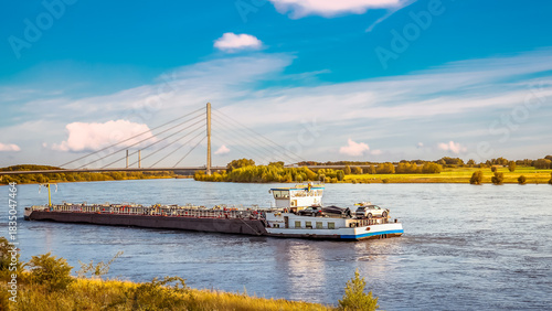 Cargo barge sailing on the Rhine River under a sunny blue sky, passing a modern cable-stayed bridge and highlighting efficient inland shipping and sustainable transport.