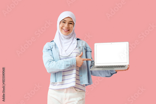 Photography Beautiful young Muslim woman in hijab pointing at modern laptop on pink backgrou