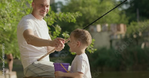 Father and son sit together by lake enjoying quiet sunny morning of fishing. Smiling caring parent removes hat of child and gently arranges strands of hair on head
