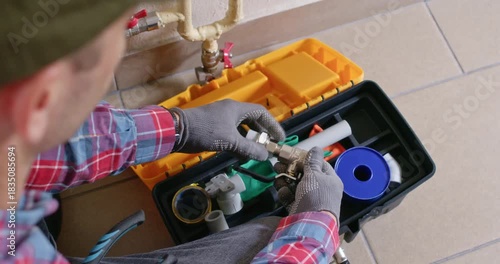 Plumber sits by toolbox selecting adapter from organized kit. Male technician reads spec sheet and matches part number preparing pipe joint for assembly