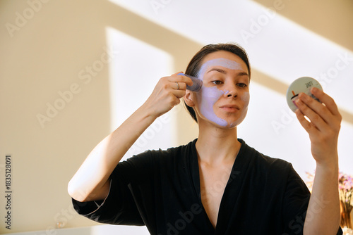 A young woman applies a clay mask in a stick to her face, looking in a mirror. Facial skin care at home