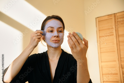 A young woman applies a clay mask in a stick to her face, looking in a mirror. Facial skin care at home