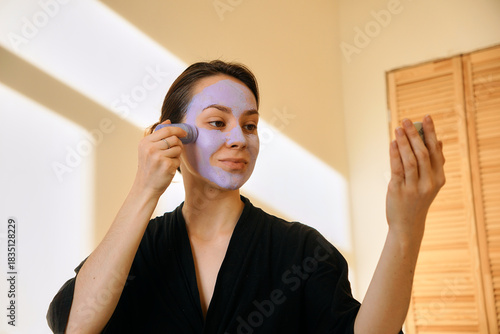 A young woman applies a clay mask in a stick to her face, looking in a mirror. Facial skin care at home