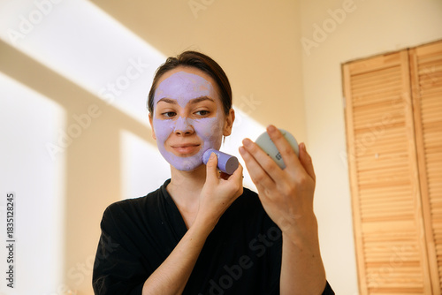 A young woman applies a clay mask in a stick to her face, looking in a mirror. Facial skin care at home