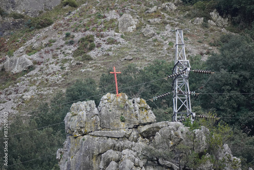 Earth dominates limestone peaks in La Hermida valley - Rural landscape featuring a wooden structure at mountain base, cross with telegraph wire and Spanish flag, set against blue sky and red earth.