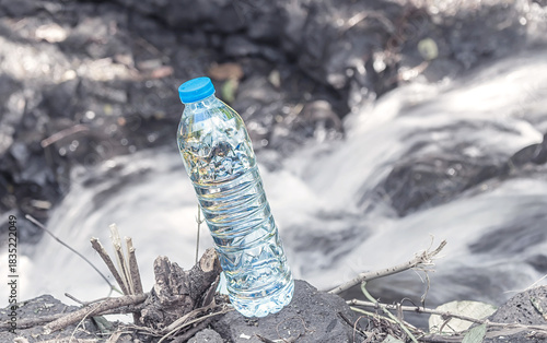A single plastic water bottle with a blue cap left on a dark rock next to a fast-flowing, blurred waterfall. Concept of plastic pollution in natural environment.