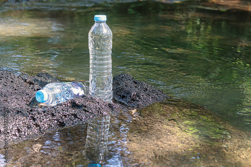 Two plastic water bottles, one standing and one lying down, abandoned on a dark, wet volcanic rock by a natural flowing stream, symbolizing environmental pollution.