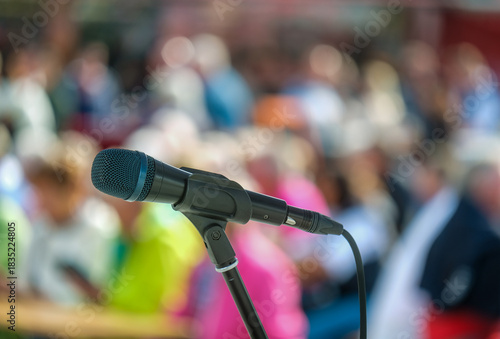Seitliche Nahaufnahme eines Mikrofons im Halter vor einer Masse an unkenntlichen Menschen im Publikum im Hintergrund auf einer Open-Air Veranstaltung – selektiver Fokus, sehr viel Tiefenunschärfe