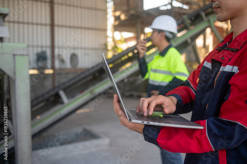 Worker Using Laptop in Industrial Setting with Colleague Talking on Walkie-Talkie in Background at Construction Site