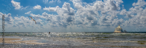 Seagull flying over ocean water near paddleboarder and old lighthouse under dramatic blue sky with copy space
