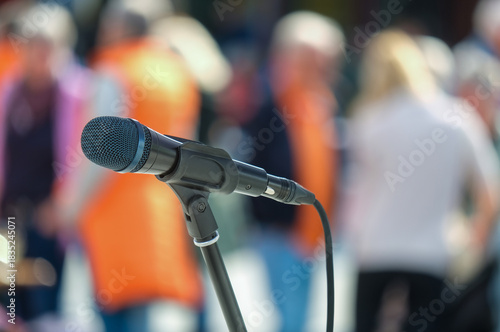 Seitliche Nahaufnahme eines Mikrofons im Halter vor einer Masse an unkenntlichen Menschen im Publikum im Hintergrund auf einer Open-Air Veranstaltung – selektiver Fokus, sehr viel Tiefenunschärfe