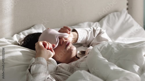 A young woman wakes up feeling unhappy, slips her sleep mask back on, and tries to fall asleep again, showing her reluctance to start the day. Concept reluctant morning.
