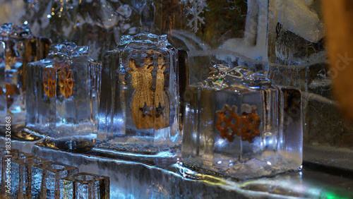 A row of crystal ice blocks on a reflective ledge showcases golden figures encased inside, glowing with blue light. A festive winter display invoking Christmas decor, art, and delicate craftsmanship.