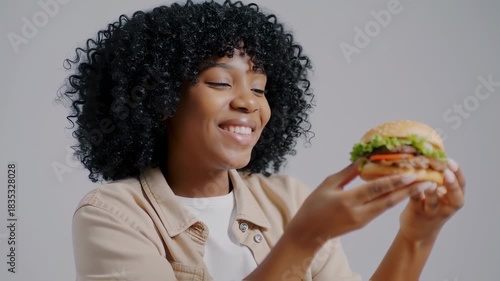 A smiling woman with closed eyes holds a burger. Curly hair, light clothing, neutral background