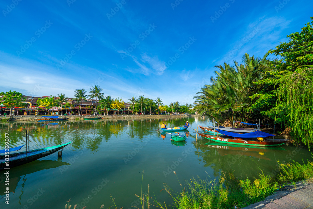 Obraz premium Wooden boats on the Thu Bon river in Hoi An ancient town. Yellow old houses on waterfront reflected in river. Hoi An is a UNESCO world heritage site in Vietnam