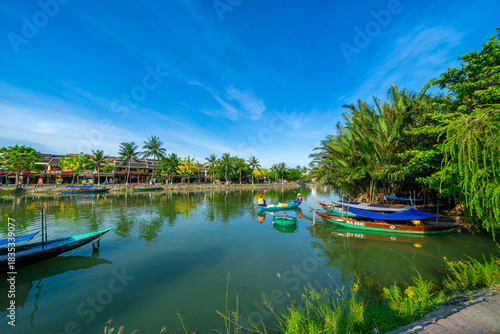Wooden boats on the Thu Bon river in Hoi An ancient town. Yellow old houses on waterfront reflected in river. Hoi An is a UNESCO world heritage site in Vietnam
