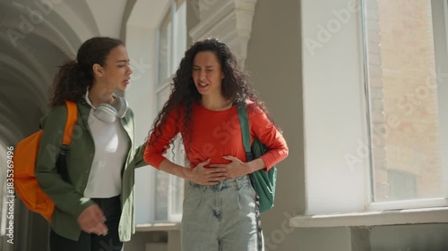 Two Hispanic Latina female students walking university girl holding stomach pain women friends in college talking feeling ache menstrual period painful cramp abdominal health problem medicine gastric